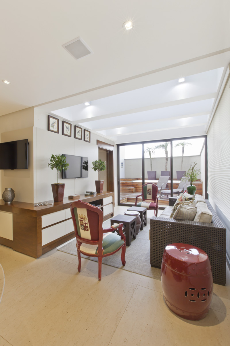 Entre o gourmet e a piscina, criou-se um ambiente de estar com TV, fechado por vidros nas laterais e teto. O móvel projetado pela profissional acompanha o desenho da parede