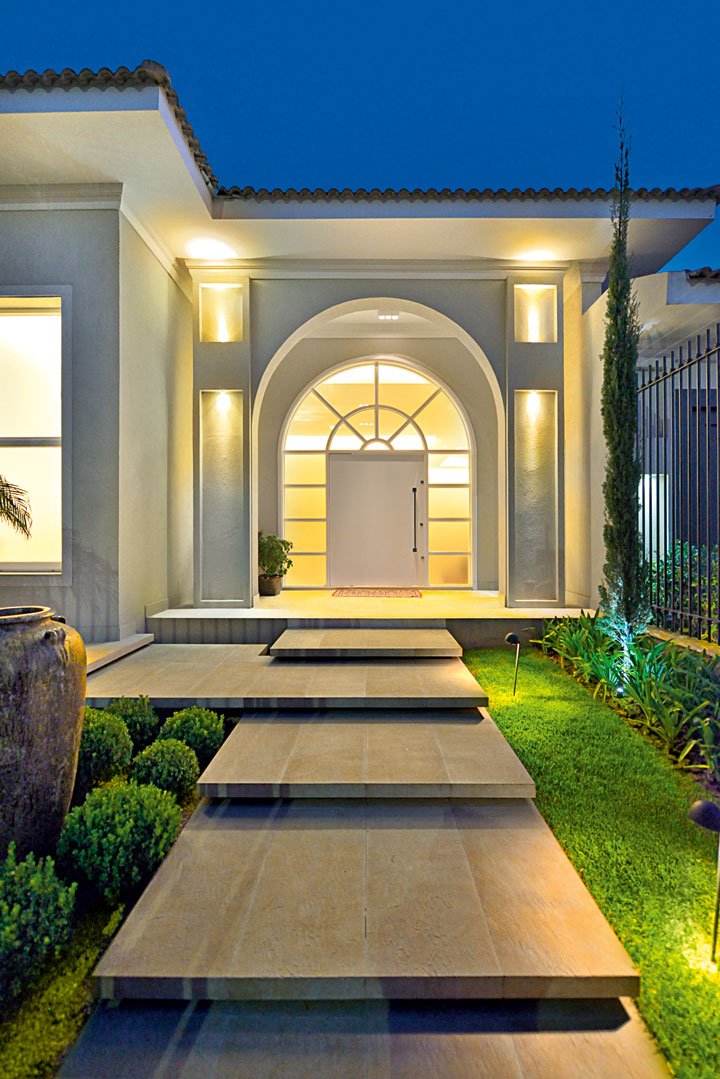 A porta curva (Marcenaria Moretti) sob o pórtico remete às linhas clássicas. Todas as esquadrias são em PVC (Shine Windows). As pisadas em porcelanato rústico da Portinari (Eco Portinari), cercadas pelo jardim, parecem flutuar e dão leveza à composição.
