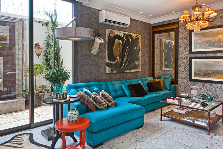 O sofá azul turquesa se destaca no home theater. A composição ficou aconchegante com a aposta nos armários cinza, papel de parede, telas e lustre clássico. O piso é em mármore Carrara. Tela do artista Sylvio Viana