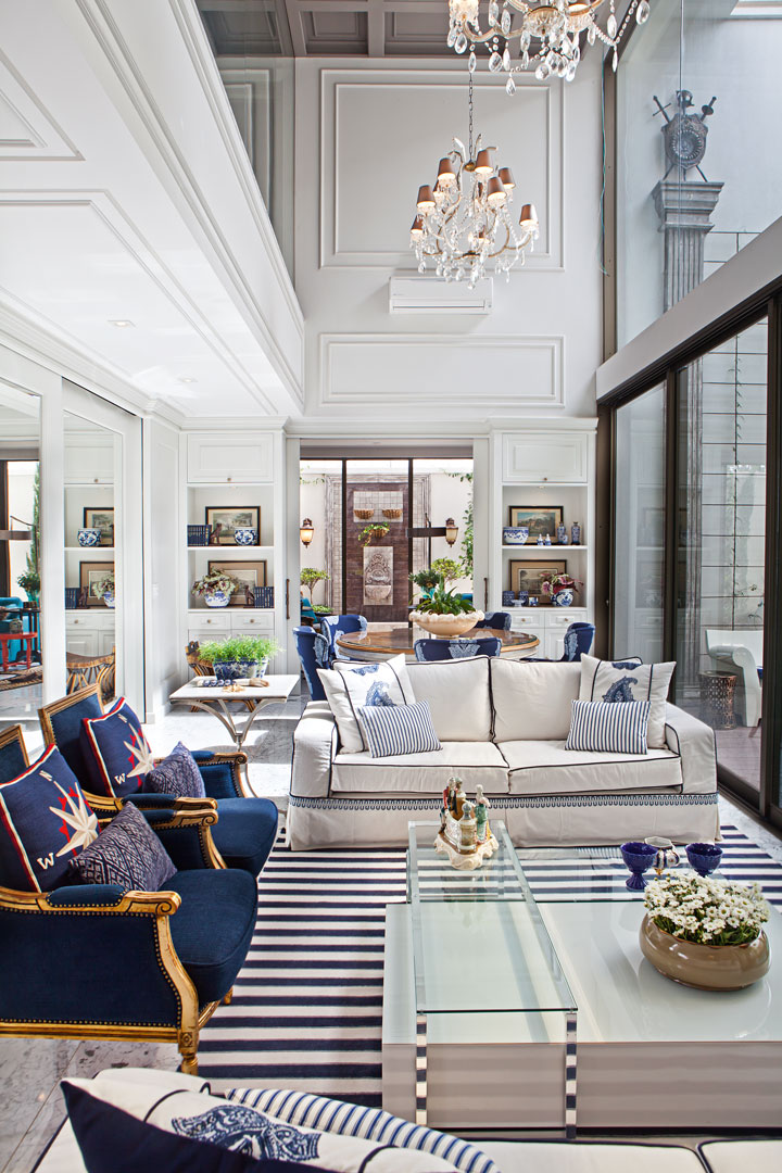 A iluminada varanda traz o charme das boiseries com a paleta navy no tapete, poltronas, mesa de centro branca (Perfil Casa) e cadeiras da mesa em madeira. Mesa lateral da Decorativa. O pano de vidro (Aluvetro) acompanha todo o pé-direito duplo e integra a piscina. Marcenaria da Gabriel e Guarinos e ar condicionado da Ar Show