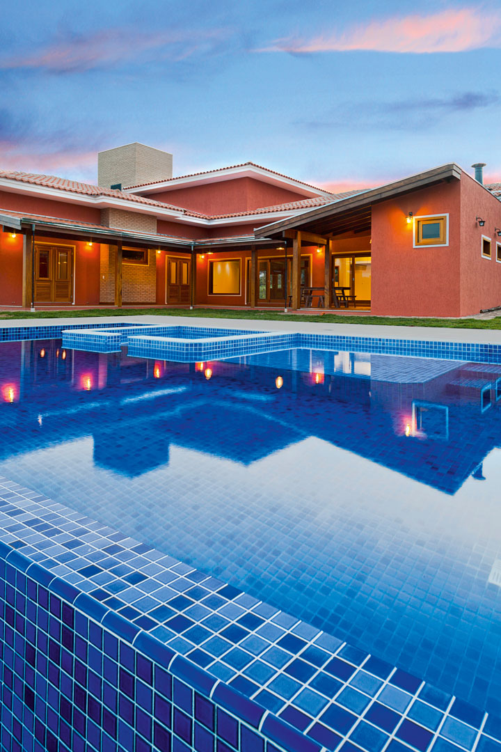A piscina com hidro e prainha tem borda infinita para acentuar a integração com o entorno. O revestimento traz pastilhas azuis da Villa Bella Acabamentos. A casa com telhas de barro e pintura em tom terroso (J. De Santis) é cercada por varandas e generosas aberturas de vidro (RN Alumínio) 
