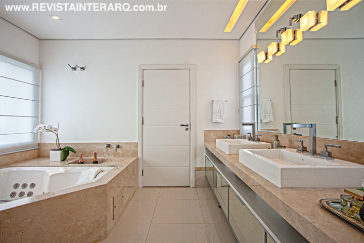 O mármore Crema Marfil foi a escolha para a bancada, banheira e detalhes da sala de banho clean do casal. Delicadamente iluminado pelas quatro janelas com persianas de tecido, o ambiente ganha ares cenográficos à noite, quando são acionadas as luzes indiretas, lâmpadas de Led ou os chuveiros com cromoterapia.
