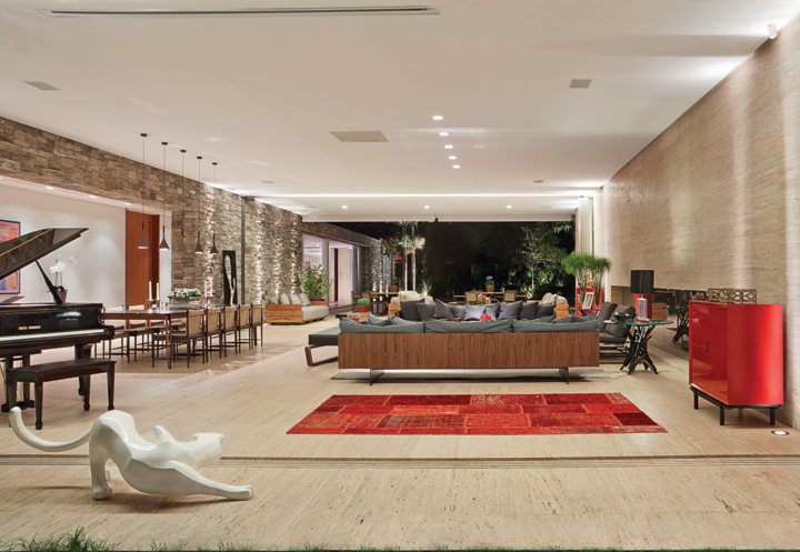 O piso em mármore Travertino bruto compõe com as pedras naturais da parede e forma o cenário ideal para ressaltar a beleza do mobiliário de design brasileiro e das peças vermelhas, como o tapete (Gallery Tapetes) e o móvel vermelho de Alessandra Delgado. A iluminação é da Light Design e a escultura “A Pantera”, da artista Vânia Barbosa. A arquitetura da casa é assinada por Valéria Gontijo
