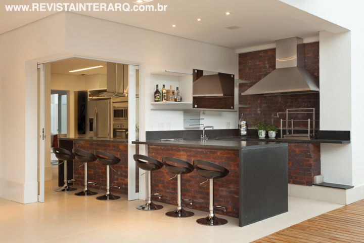 A cozinha e o espaço gourmet são separados apenas por uma porta de vidro que, quando aberta, integra totalmente os ambientes. A unidade visual é acentuada pela bancada em tijolos da Palimanan e tampo em granito