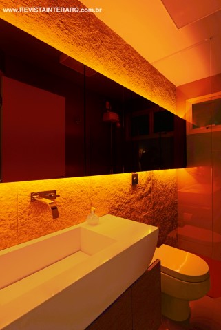 O banheiro social foi elaborado com materiais para trazer modernidade e relaxamento. As paredes ganharam revestimentos em pedras e placas de alumínio na cor fendi. A bancada “canoa” em Corian (Granipedras) e um grande armário (Wood Design) com espelhos (Transparência Design de Vidros) foram valorizados pela iluminação (Ilumax) 