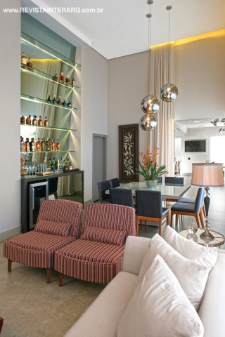 Móveis de linhas retas, como as poltronas da Visual Q, ambientam o living. O jantar foi setorizado pelos pendentes Bola Prata (NR Iluminação) e pelo arrojado bar com espelho fumê inclinado e Led (execução da Decora Móveis), criado para acomodar a adega e coleções de garrafas do morador. Todas as cortinas da casa são da Nice Decorações. O piso é em porcelanato concreto escuro da Portinari 