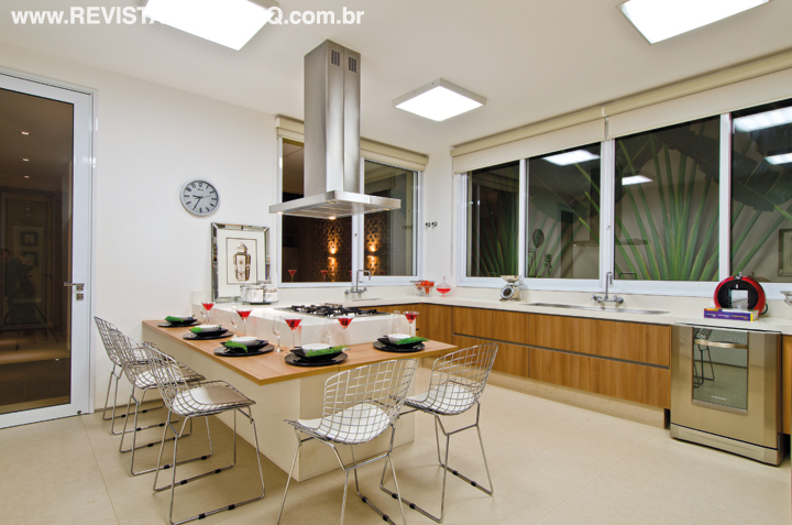Ilha com cooktop e bancada para lanches rápidos em madeira e Caesarstone Snow (Projeto de Pedra) revelam a funcionalidade da cozinha. Os armários amadeirados são da Marcato