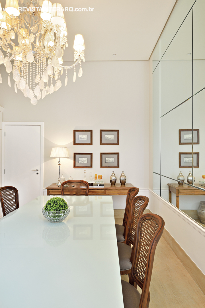 As cadeiras em madeira com encosto em palhinha contrastam com a mesa em laca branca do jantar. A parede com espelhos bisotês duplicam a beleza do lustre de cristais de rocha. O piso de todo o social foi repaginado com mármore Travertino Perlato