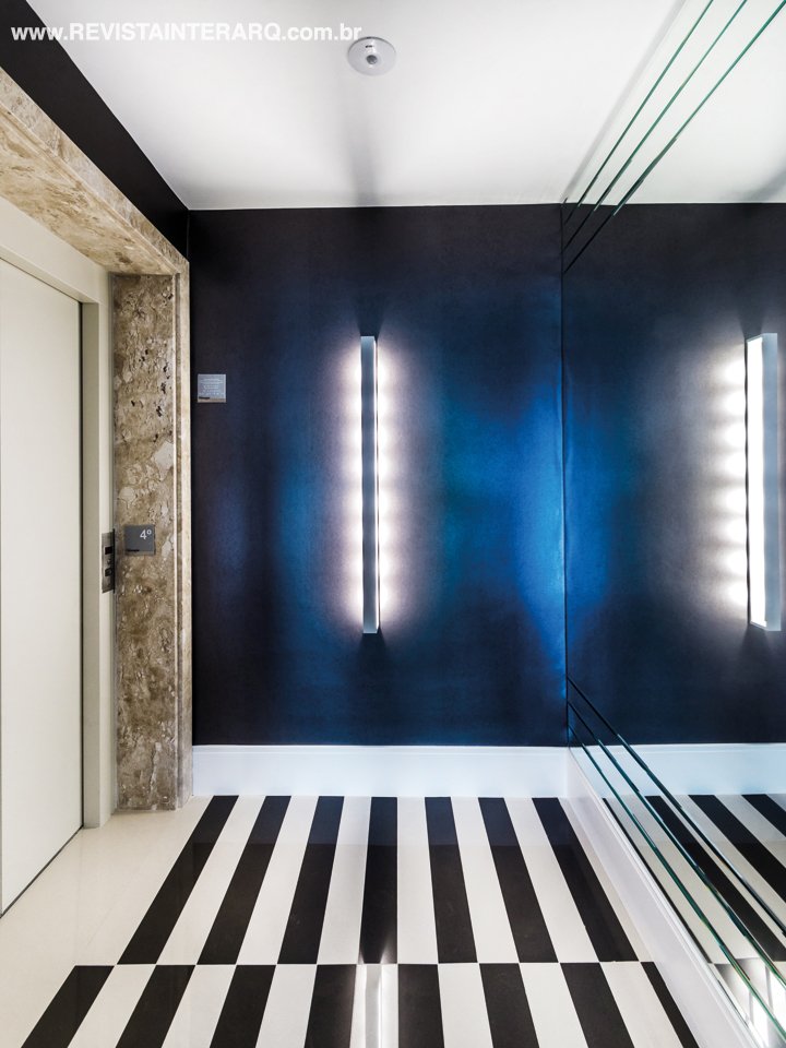 A paginação gráfica do piso em porcelanato grafite e off-white (Toque Final) recebe, com estilo, quem chega ao hall principal. Na parede, a ousadia ficou por conta do revestimento escuro da Orlean com a arandela vertical (Light Concept)