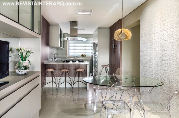 A moderna cozinha traz banquetas para lanches rápidos e mesa de vidro com cadeiras de acrílico (Firmato Móveis) para dar leveza ao ambiente. O revestimento em alto relevo é da Decortiles (Toque Final) e a luminária é da Light Concept