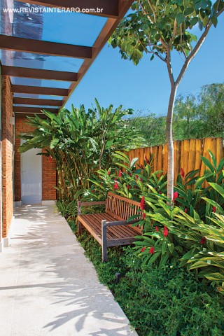 Unido visualmente ao living, o jardim com Alpínias, Agapantos, Helicônias e Cambará rasteiro conta com um banco de madeira de demolição, posicionado à sombra de um Ipê