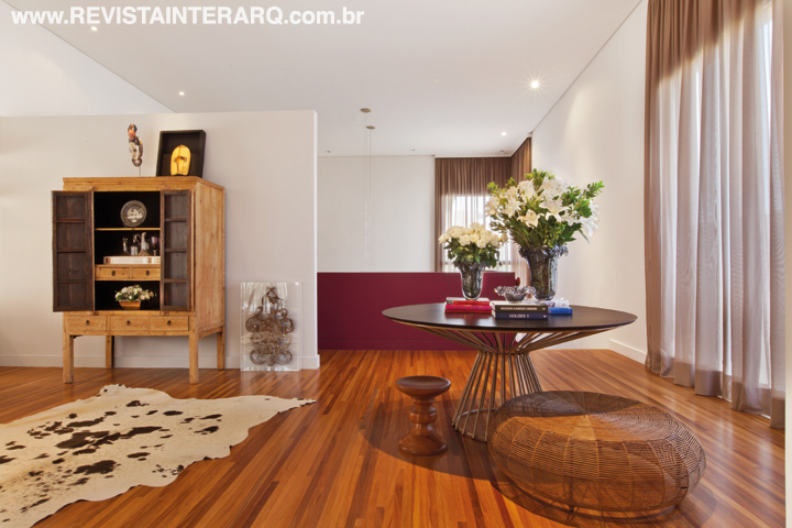Bonito e acolhedor, o piso em madeira Cumaru foi colocado por cima do porcelanato existente na casa. No hall de entrada, uma parede de gesso pintada de cinza “esconde” o corredor dos quartos. O guarda-corpo da escada de acesso à sala de jantar era de vidro e foi substituído por um em gesso pintado de cor forte. O décor mescla armário tailandês, tapete de pele de vaca, pufe de fibra natural, Muranos, mesa redonda e um original fragmento de madeira em caixa de acrílico