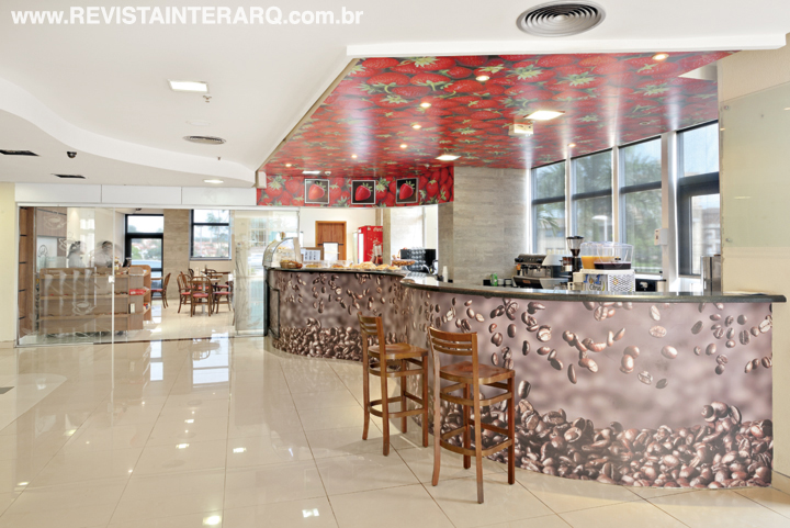 O balcão de linhas orgânicas e imagem com grãos de café convida a uma pausa para degustar a bebida. No teto, o tom quente dos morangos atrai o olhar e anuncia um cardápio eclético
