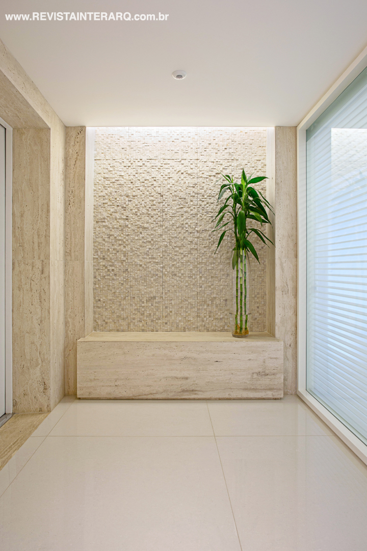 No hall de entrada, banco e molduras em mármore Travertino Romano bruto destacam a parede com mosaico em mármore rústico