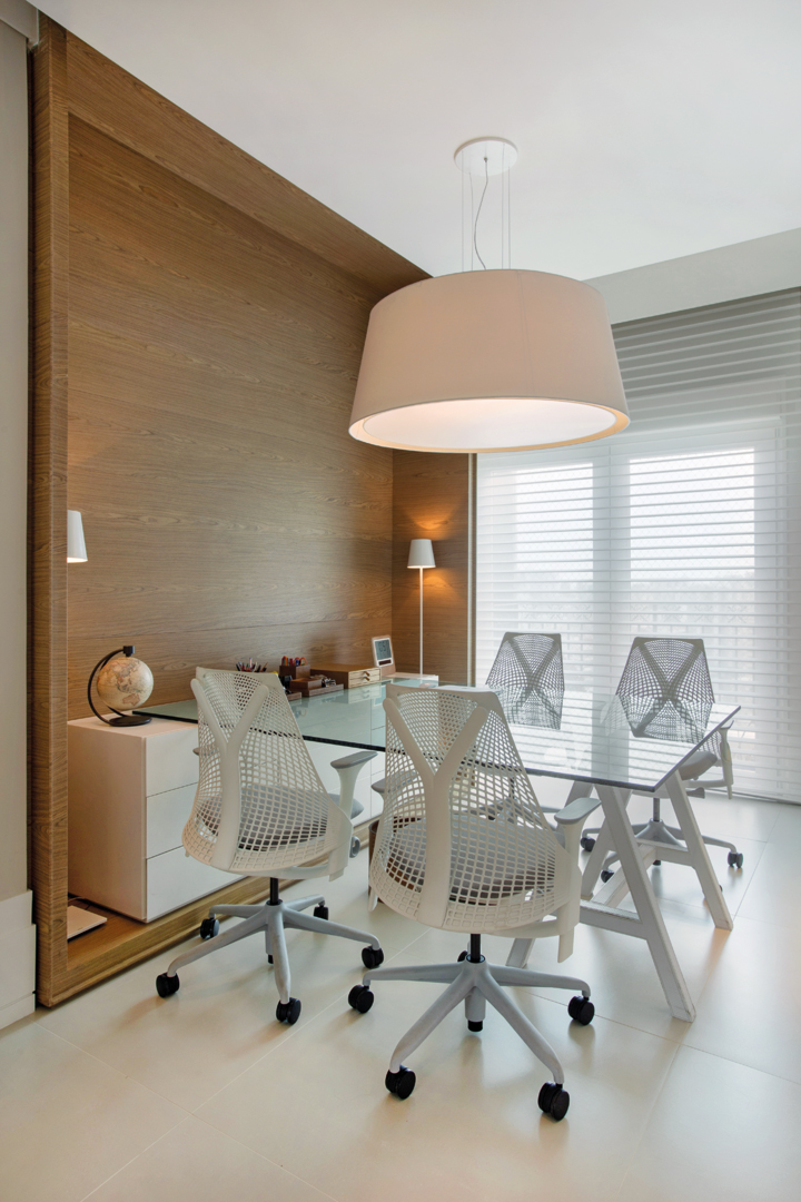O office conta com uma mesa de reuniões feita com tampo de vidro apoiado no móvel planejado (Incomap Móveis) e em um cavalete todo revestido em couro pespontado (Le Cadeau Curtume Casa). Cadeiras Sayl, persianas Hunter Douglas Luxaflex (Délicatesse Tecidos) e pendente da Kelvin LD