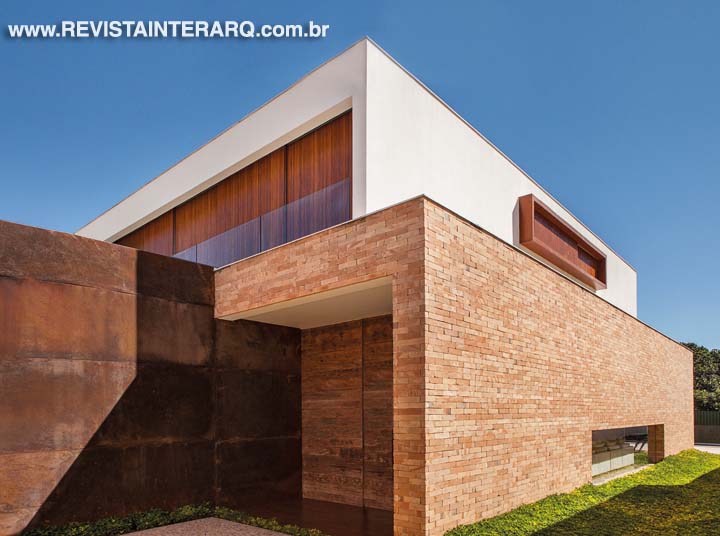 Um grande painel em aço corten acompanha toda a entrada da casa, no alto, com porta principal em madeira de demolição.