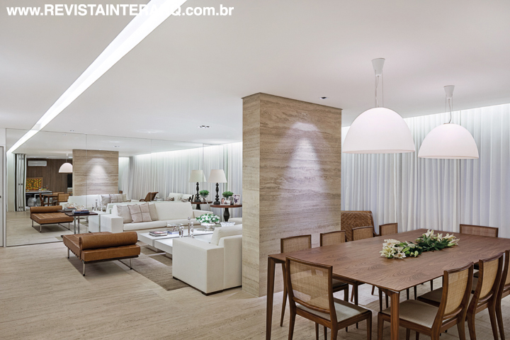 O painel espelhado cria um efeito lúdico, duplicando o ambiente social, revestido com mármore Travertino Romano Bruto (Marmoraria Alvorada). Os estofados off white da Artefacto, elegantemente emoldurados pelo tapete da Gallery Tapetes ganham aconchego quando combinados com a madeira e com o couro natural do recamier Barcelona, desenhado por Mies Van der Rohe. Na sala de jantar, pendentes Mamma