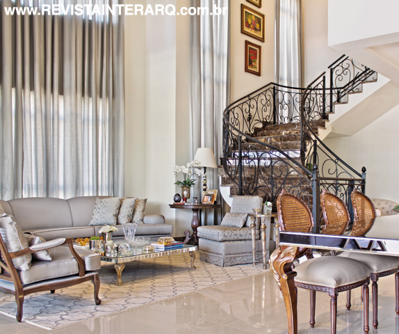 Cortinas de linho acompanham o pé-direito duplo do social, que tem como protagonista a escada com degraus em mármore marrom e corrimão de ferro trabalhado. O living com sofás cinzas (Estofados Cardoso) e poltrona clássica (Sierra Perfil Casa) é delimitado pelo tapete claro sobre o piso em porcelanato
