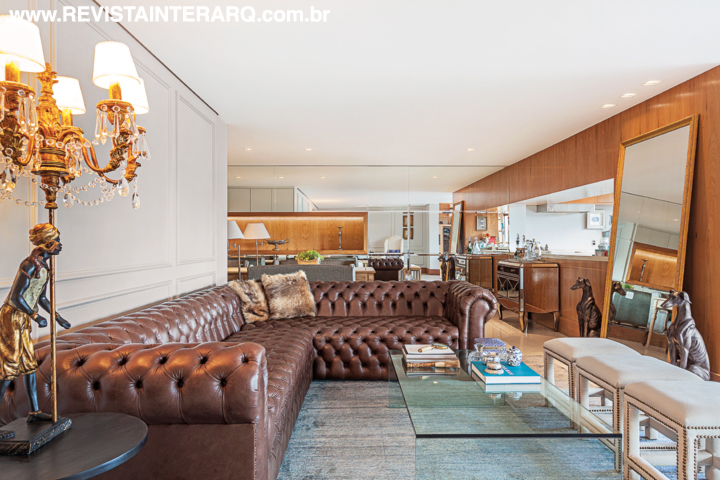 O sofá Chesterfield em couro marrom (Estofados Cardoso) e o tapete da By Kamy delimitam o espaço do living, decorado ainda com mesa de vidro e almofadas de pele. Ao fundo, a parede espelhada duplica visualmente o espaço