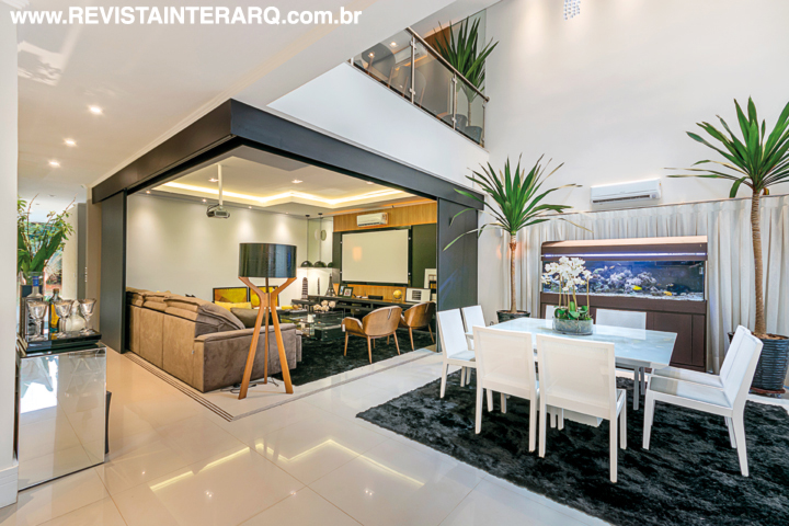 O home theater com móvel da Novo Conceito Móveis e estofados confortáveis pode ser totalmente integrado com a sala de jantar e a cozinha. o jantar conta com mesa branca valorizada pelo tapete escuro 