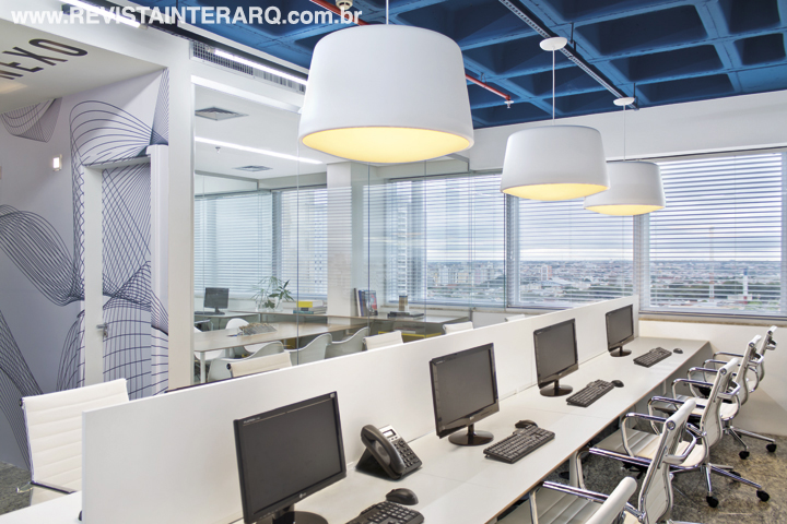 Nas estações de trabalho, o forro aparente foi pintado de azul. Luminárias pendentes (Lumilight) acompanham a mesa de trabalho, que também recebe muita luz natural graças às grandes janelas com persianas