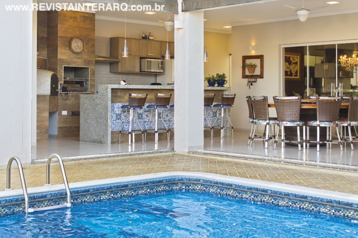 A varanda em “L” abriga mesa de refeições com cadeiras em fibra sintética (Espaço Fibras Decor Home) e área da churrasqueira, revestida com porcelanato com estampas de madeira e de azulejos portugueses (Casa dos Construtores)
