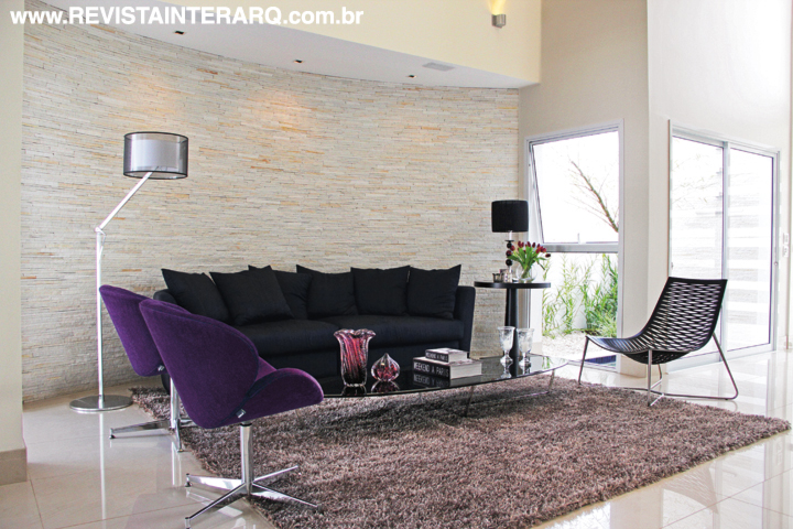 A luz natural invade os ambientes internos, delineando as peças do decor, como as poltronas roxas e o sofá preto (tudo da Robusti) do living. Luminária de piso da Luminá Iluminação. Cortinas da ACB Casa e acessórios da UneRobusti