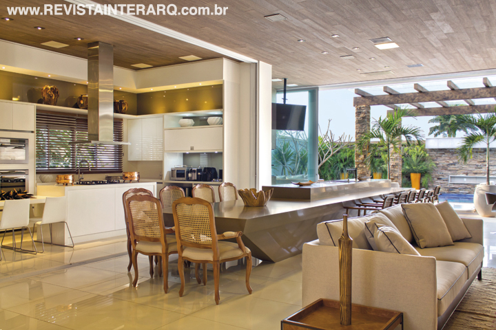 A cozinha com armários da Kitchens se abre totalmente para a varanda contemporânea, ambientada com uma arrojada bancada em Limestone (Stone Center) em contraponto às cadeiras clássicas de madeira e palhinha (Perfil Casa). O forro confere aconchego com o porcelanato amadeirado da Casa dos Construtores. Toda iluminação conta com luzes de Led, da ViaLight. Coifa e balcão refrigerado da Refrigeração Araçatuba. Os grandes vãos de janelas e portas receberam esquadrias de alumínio e vidros da Aluvetro. O projeto arquitetônico é de Fabiano Hayasaki