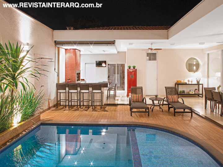 A piscina com pastilhas de porcelana azul contrasta, com o entorno em porcelanato amadeirado da Linha Ecowood (Portobello Shop) e com o pequeno jardim com espécies tropicais. Pastilhas brancas delimitam a área do chuveirão, enquanto vermelhas, em contraste com as parede claras (Tintas e Texturas Maggicor), ressaltam a churrasqueira. Os móveis são da Copav e a bancada em granito Marrom Café foi executada pela Marmoraria Exclusive. 