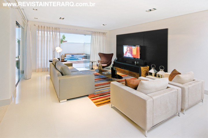 O living cumpre dupla função neste projeto e integra sala de estar e TV. A ambientação contemporânea fica por conta do estofado em suede (UneRobusti) e painel em madeira ebanizada feito sob medida. O tapete colorido foi confeccionado na Índia com exclusividade para esta residência
