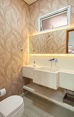 O papel de parede (Magnificat) confere movimento ao lavabo e acompanha o tom da bancada em Silestone Blanc Leather. A iluminação atrás do espelho cria um efeito interessante