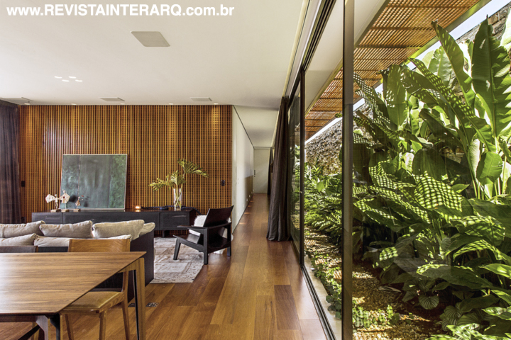 No social, a transparência das portas de correr (Esquadrias 2 irmãos) inclui o exuberante jardim lateral no décor com móveis de design e luminotécnica automatizada (Studio Q). Forro de gesso da Abece Gesso. Os materiais usados, como madeira, concreto, pedra e aço corten, não apenas determinam o acabamento como também os usos específicos