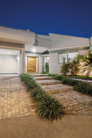No paisagismo que envolve a construção, a arquiteta trabalhou com diferentes texturas e cores de verde. Na fachada, os jardins complementam os caminhos de pedras Quadrato Tropical amarelo (Villa Bella Acabamentos)