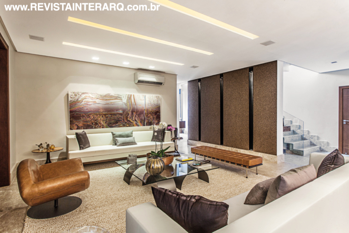 A parede revestida com papel de parede Mica marrom (Zuhause Dekor) delineia o limite entre o living e a escada. A ambientação traz móveis de linhas retas e peças de design, como a poltrona Gaivota e a mesa de centro Arcos, de Ricardo Fasanello (tudo da Arquivo Contemporâneo)