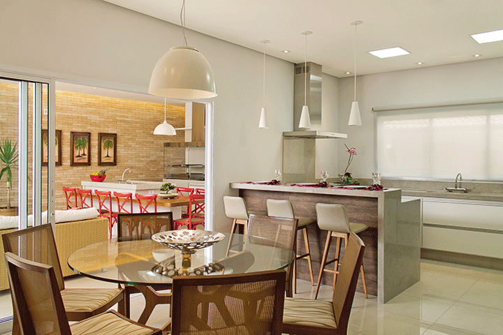 Interligada ao lazer e à sala de almoço, a cozinha tem conceito aberto e moderno. Os diferentes ambientes são marcados pelos pendentes brancos. A mesa de refeições e as banquetas são da Decorativa e coifa é da Refrigeração Araçatuba
