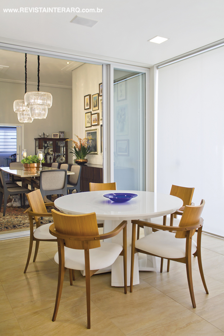 Junto à ala social, a mesa em laca branca é mais uma opção para acomodar família e amigos na varanda. O piso é em porcelanato Navona Crema, da Portobello Shop e o prato azul é da Vera Orsi. Persiana rolô da Griffe A. O projeto original da casa é de Leder Ferreira, com intervenções de Suelen Fornazari e Giuliana Jacob