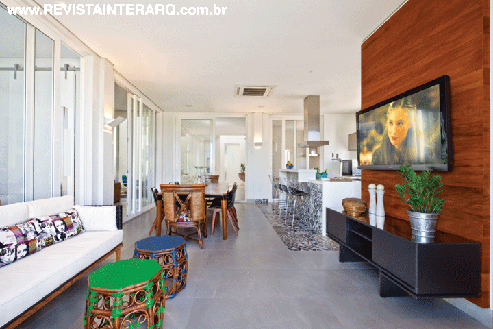 Receptivo, o estar da varanda ganhou móveis de fibra e madeira (Artenal), almofadas estampadas e cortinas de linho (Tenda Decorações). O painel de madeira com estante preta permite que a TV possa ser vista também da mesa de jantar (Perfil Casa), posicionada em frente à bancada da área gourmet. A coifa é da Refrigeração Araçatuba