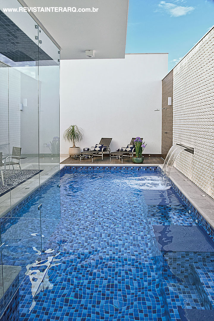 A piscina em forma de raia se estende entre as áreas externa e interna, passando pela sauna envidraçada. As prainhas em Silestone (Fran Mármore) ocupam uma das laterais, onde um pequeno véu de água jorra da cascata incrustada na parede