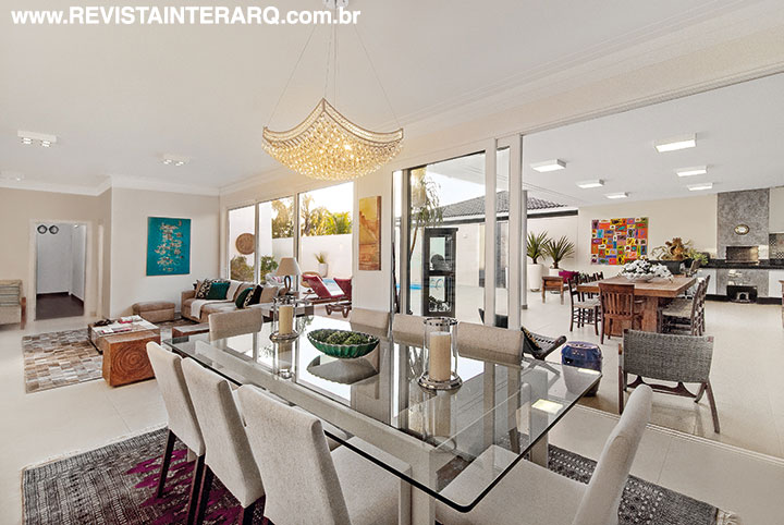 O design moderno do lustre de cristais (Reflexo Iluminação) acompanha a personalidade da mesa com tampo de vidro e cadeiras neutras. Abertas, as portas de PVC da FScott integram a área de lazer