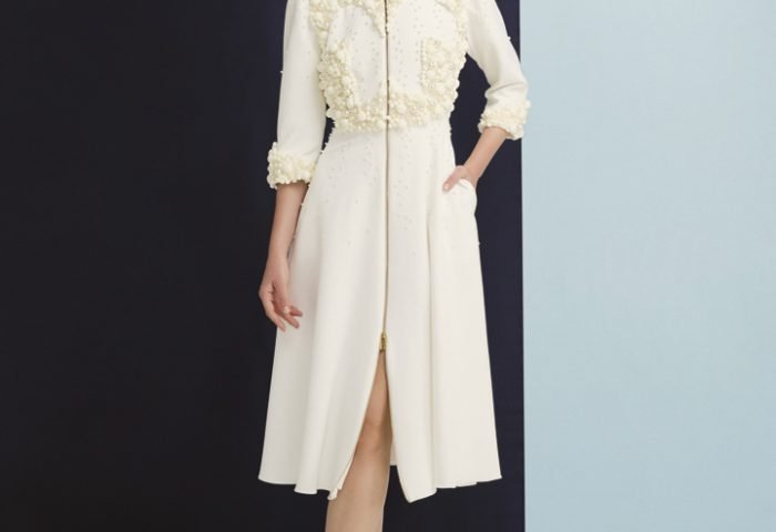 O vestido com abertura na frente em cor bege apresenta delicadas aplicações de pérolas e pompons na parte frontal e mangas.