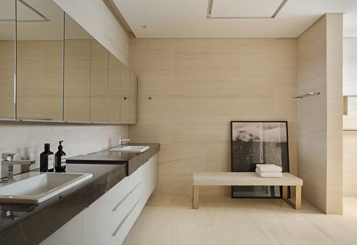 A sala de banho conta com bancada em mármore Gris Armani (Stone Center com execução Granitex) e armários (Kitchens) para ampliar o ambiente. Banco e quadro da Jackie Móveis. Iluminação pontual da Lumilight. O mesmo Limestone foi aplicado no chão e nas paredes